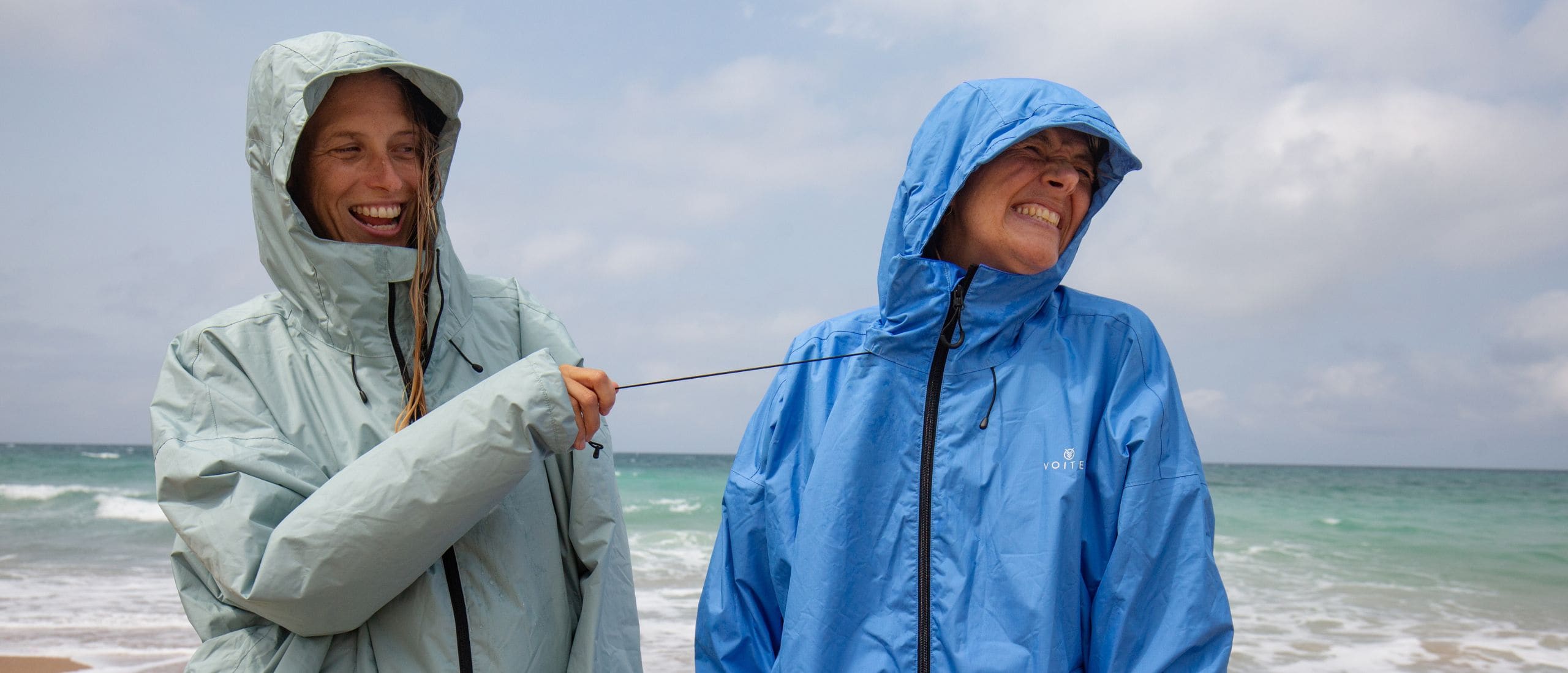 Two people wearing lightweight hooded rain jackets - All weather Coat stand on a beach, laughing while one pulls on the drawcord of the other jacket. The jacket on the left is a pale mint green and the jacket on the right is a medium blue.