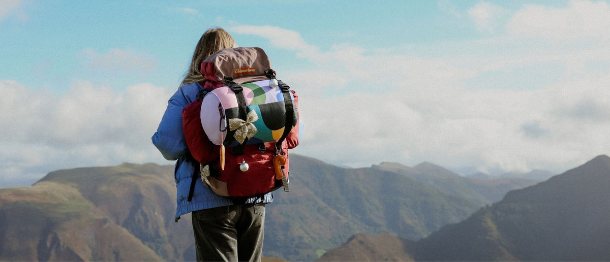 VOITED Outlet: hiker seen from behind with a backpack and a colorful VOITED blanket strapped on, decorated with a festive bow, overlooking green mountains.