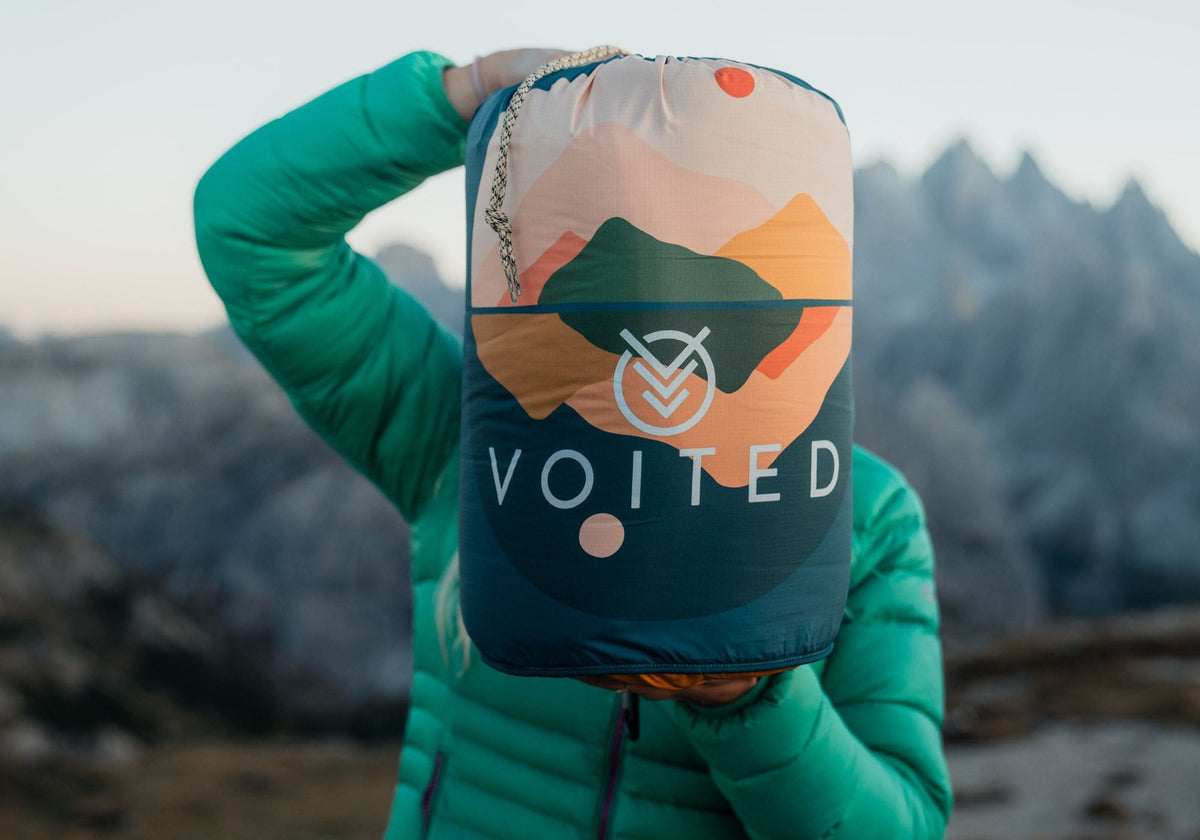 A person wearing a bright green puffer jacket holds up a Voited backpacking bag outdoors in front of a blurred mountain landscape. The bag has a colorful mountain graphic and the brand name 'VOITED' printed on the front.Cloudtouch blanket

