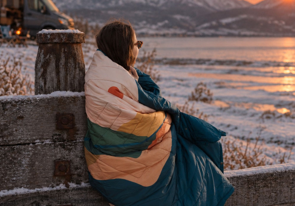Woman wrapped in a VOITED Cloudtouch blanket with a colorful peach, mustard and green pattern, looking out over a snowy lake at sunset. AI modified