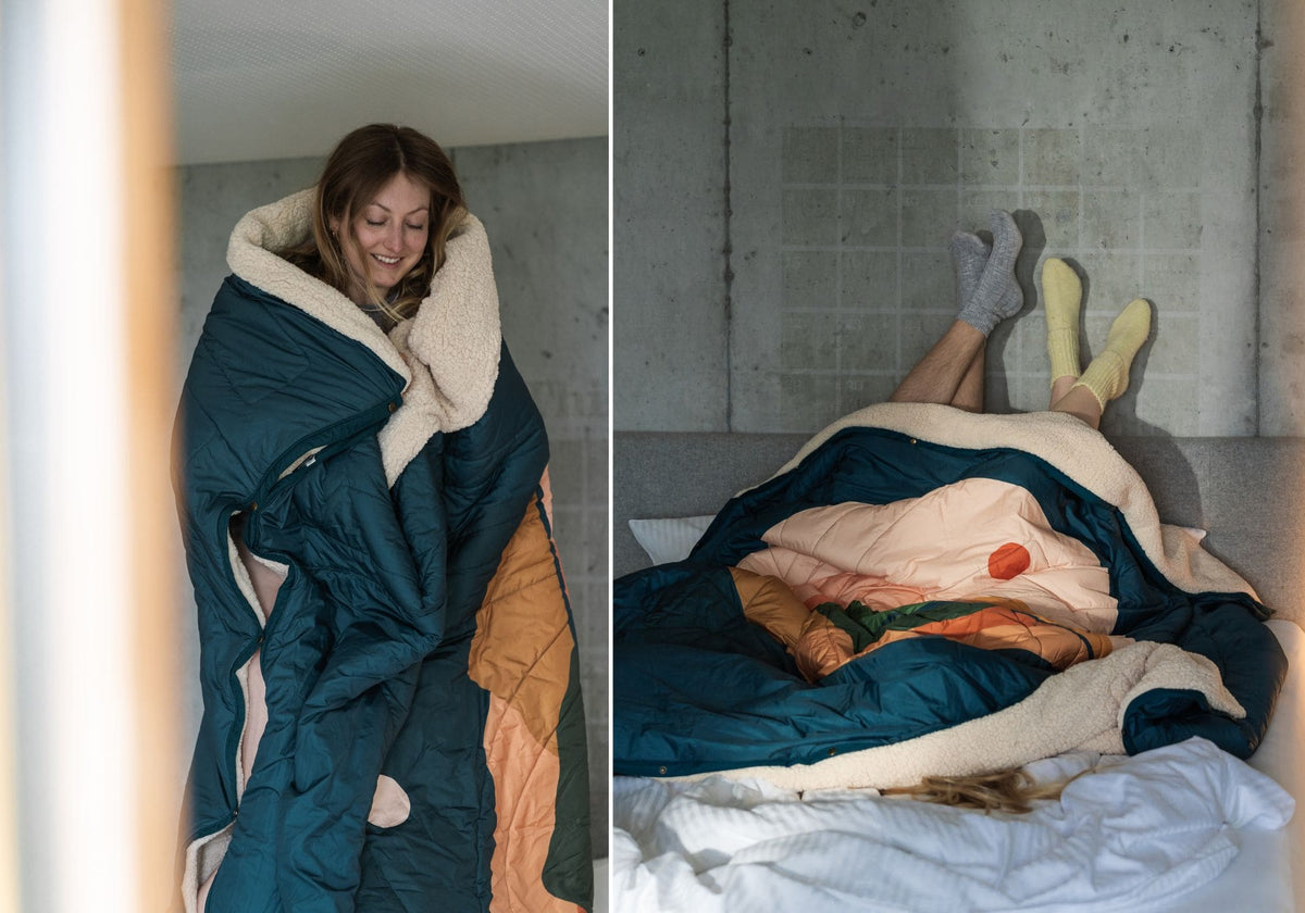 Woman wrapped in a VOITED Cloudtouch  QUEEN blanket with cozy sherpa lining; two people lying under the blanket on a bed with their socked feet up against a concrete wall.