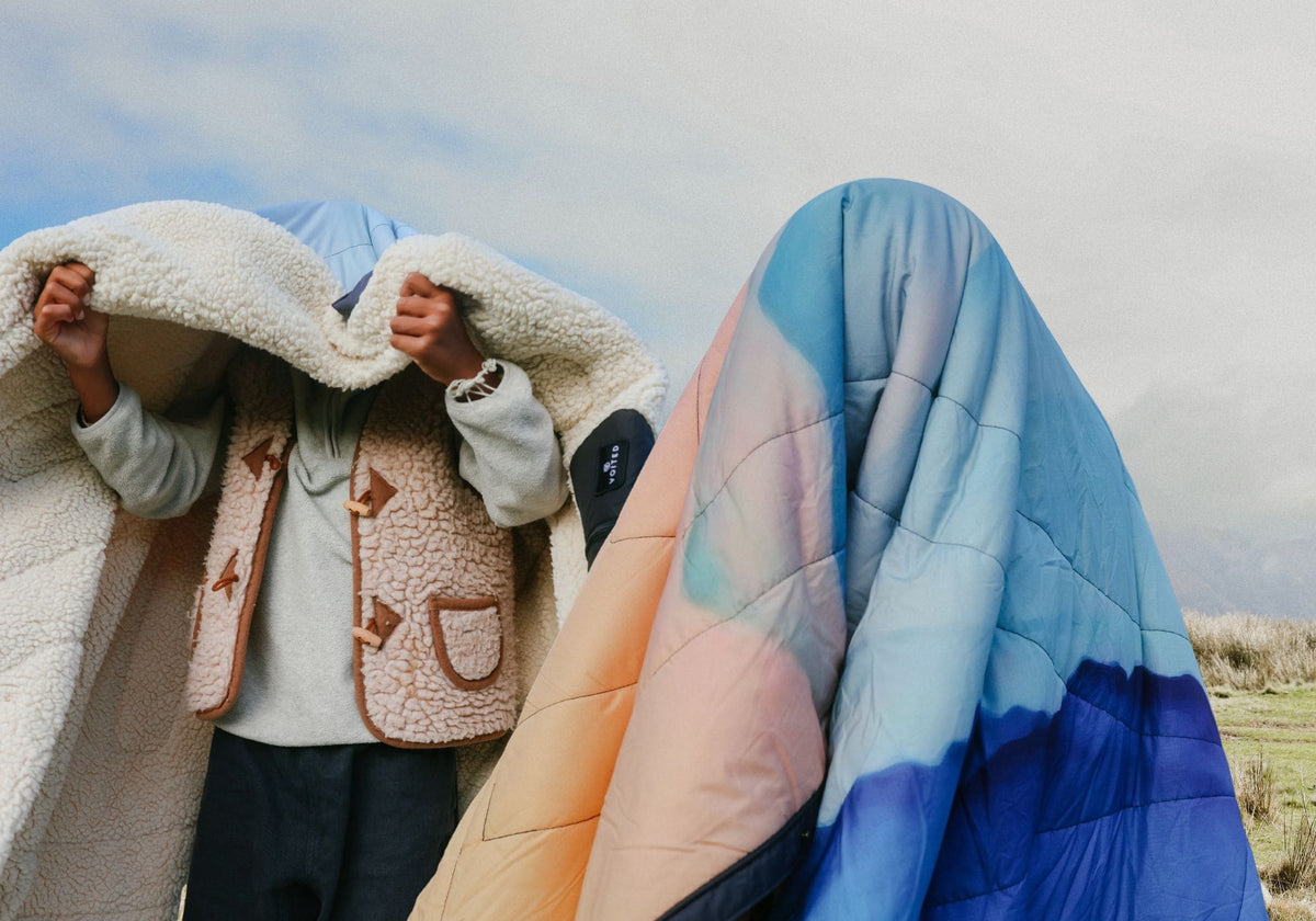 Two people outdoors under a cloudy sky. One wraps themselves in a fluffy sherpa blanket, the other is fully covered by a quilted Voited Cloudtouch blanket with a gradient from peach to light blue to deep blue.