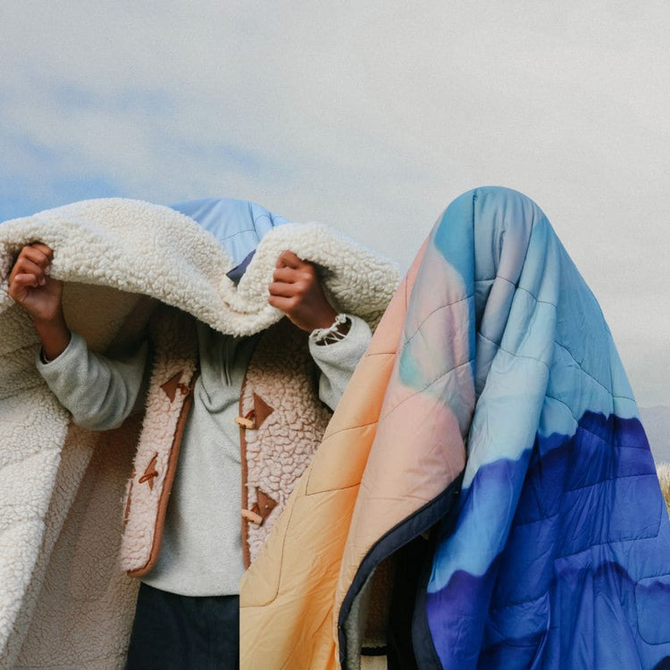 Two people outdoors under a cloudy sky. One wraps themselves in a fluffy sherpa blanket, the other is fully covered by a quilted Voited Cloudtouch blanket with a gradient from peach to light blue to deep blue.