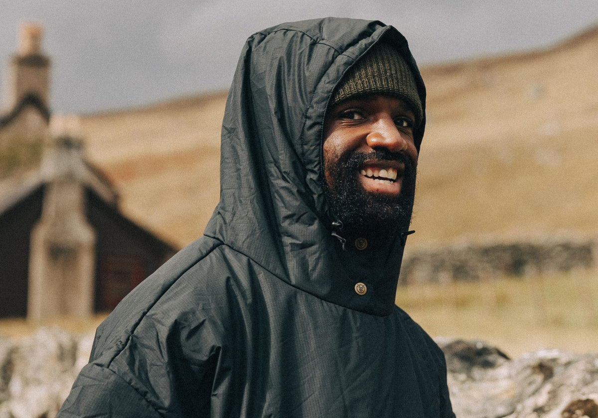 A smiling man wearing a dark green VOITED poncho with hood up over a beanie, standing outdoors in front of a rolling hillside and a stone cottage.