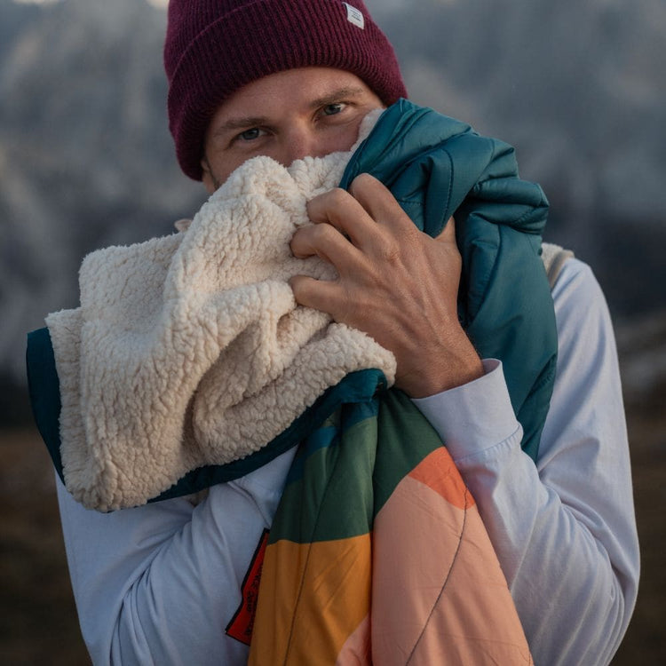 A man outdoors in a mountain setting, wearing a burgundy beanie, holds up a VOITED Cloudtouch Blanket with a cream sherpa fleece side and a teal and orange side, with mountains blurred in the background."