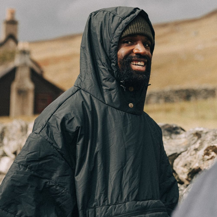 A smiling man wearing a dark green VOITED poncho with hood up over a beanie, standing outdoors in front of a rolling hillside and a stone cottage.