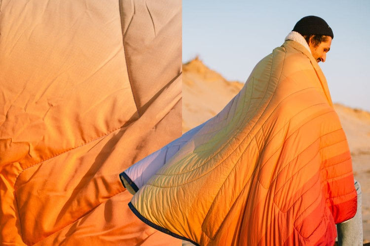 A quilted puffer blanket with a soft white sherpa collar, shown in a close-up detail view on the left and worn by a person in a desert setting on the right. The blanket features a vibrant gradient pattern transitioning from orange and yellow to light blue and purple. CloudTouch® Blanket
