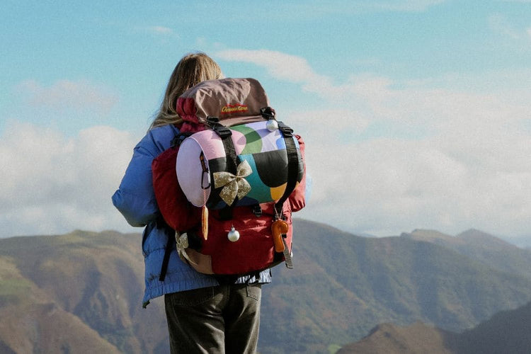 VOITED Outlet: hiker seen from behind with a backpack and a colorful VOITED blanket strapped on, decorated with a festive bow, overlooking green mountains.