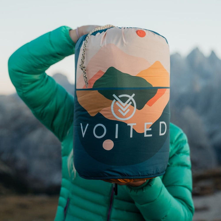 A person wearing a bright green puffer jacket holds up a Voited backpacking bag outdoors in front of a blurred mountain landscape. The bag has a colorful mountain graphic and the brand name 'VOITED' printed on the front.Cloudtouch blanket
